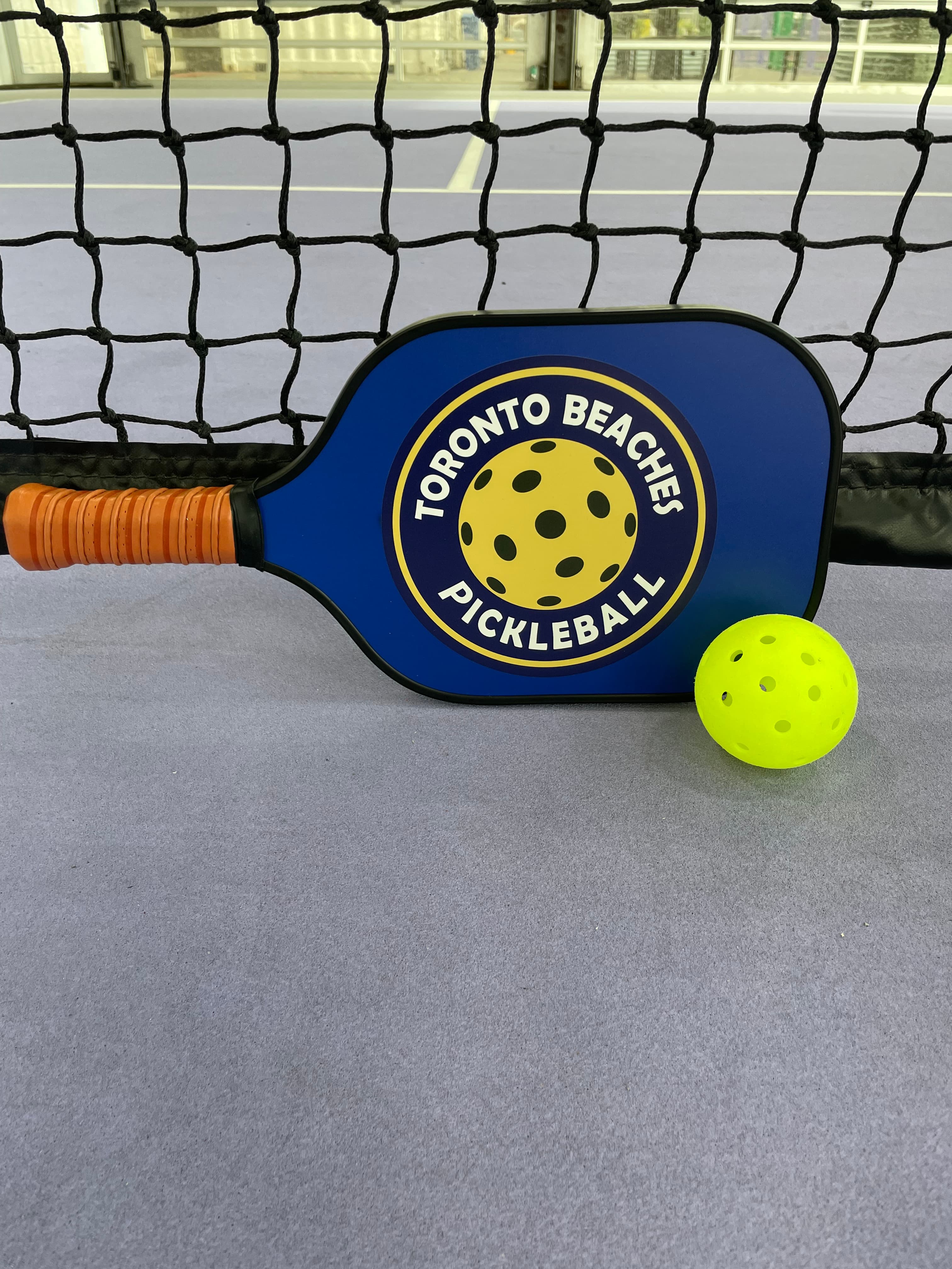 Blue Toronto Beaches Pickleball paddle and yellow ball leaning against a black court net.